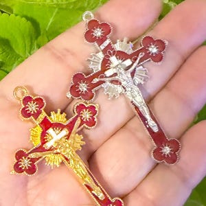 May include: Two ornate crucifix pendants. One is gold-toned with a red enamel cross and a white Christ figure. The other is silver-toned with a red enamel cross and a silver Christ figure. Both have floral accents and are held in a hand.