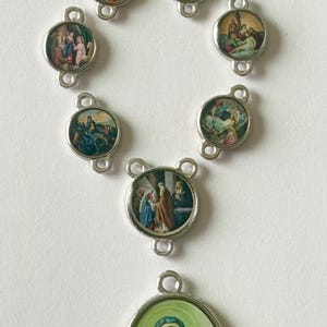 Pu&ograve; includere: Un rosario religioso con maglie in metallo argentato e ciondoli circolari. Ogni ciondolo presenta un'immagine colorata e dettagliata che raffigura scene della Bibbia. Il ciondolo inferiore mostra un ritratto di donna con un velo blu.