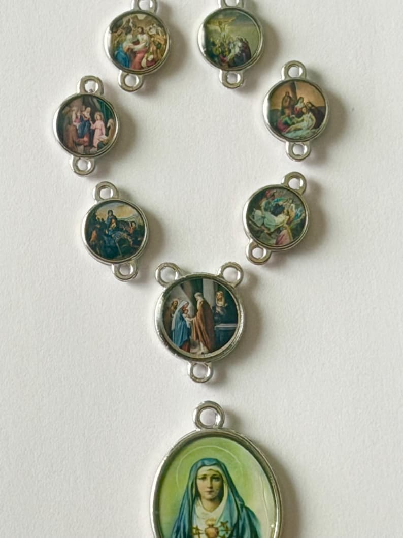 Pu&ograve; includere: Un rosario religioso con maglie in metallo argentato e ciondoli circolari. Ogni ciondolo presenta un'immagine colorata e dettagliata che raffigura scene della Bibbia. Il ciondolo inferiore mostra un ritratto di donna con un velo blu.