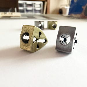 May include: Close-up of several metal beads with cross-shaped cutouts. The beads are in various colors, including gold, silver, and a muted green. The beads are cube-shaped with a hole through the center.