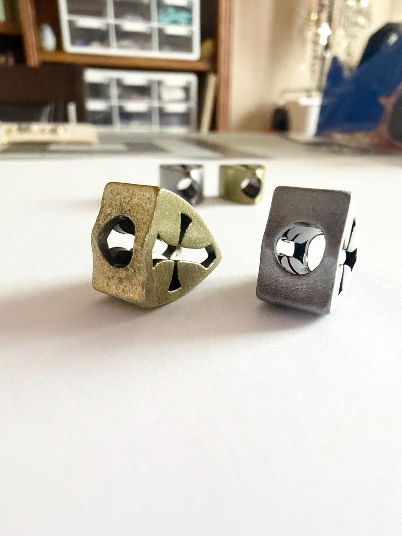 May include: Close-up of several metal beads with cross-shaped cutouts. The beads are in various colors, including gold, silver, and a muted green. The beads are cube-shaped with a hole through the center.