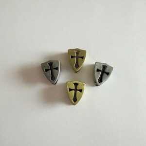 May include: Four metal shield charms with a cross design. Two charms are silver, and two are gold. The charms are arranged in a circular pattern against a white background. The charms are likely for jewelry making or crafting.