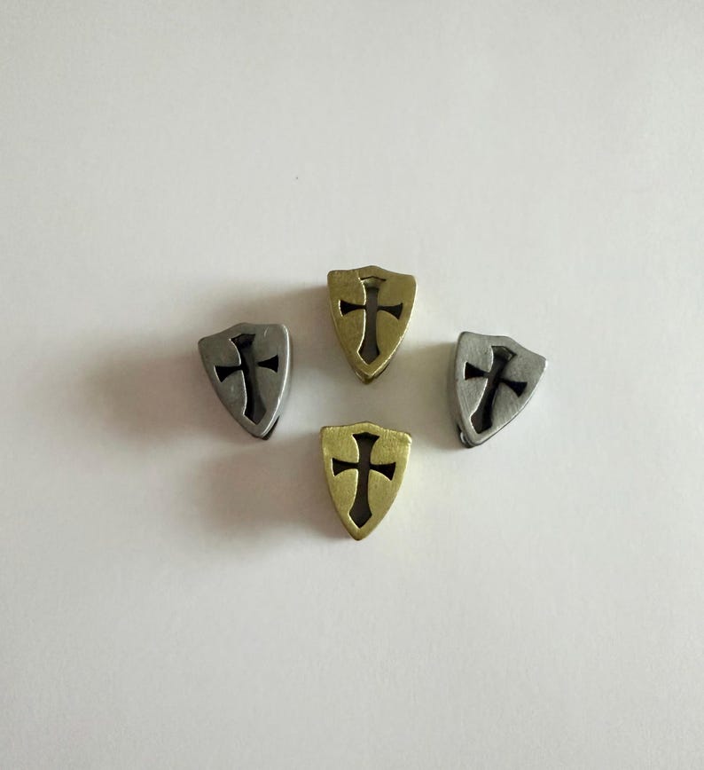May include: Four metal shield charms with a cross design. Two charms are silver, and two are gold. The charms are arranged in a circular pattern against a white background. The charms are likely for jewelry making or crafting.