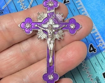 May include: A purple and silver crucifix pendant. The cross features a silver Jesus figure, silver sunburst accents, and four decorative flower-shaped accents with clear rhinestones. The pendant is held against a blue background.