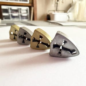 May include: Four metal stud earrings in a row, each with a cross cutout. The earrings vary in color, including gold, silver, and a darker metallic hue. The earrings are on a white surface.