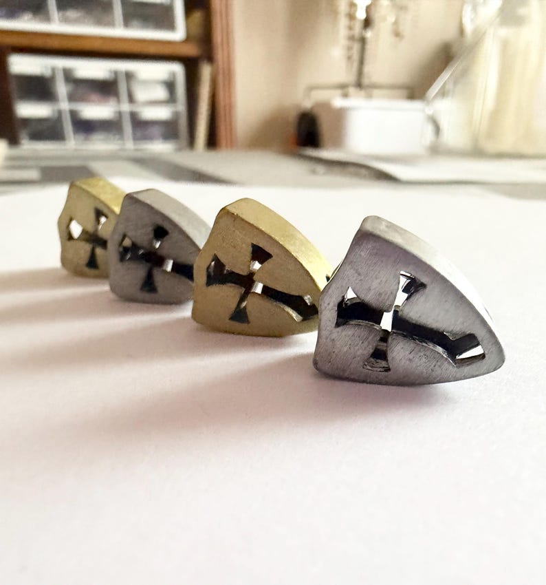May include: Four metal stud earrings in a row, each with a cross cutout. The earrings vary in color, including gold, silver, and a darker metallic hue. The earrings are on a white surface.