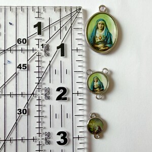Pu&ograve; includere: Tre ciondoli religiosi con montatura color argento. Il pi&ugrave; grande &egrave; ovale, quello centrale &egrave; rotondo con tre anelli e il pi&ugrave; piccolo &egrave; rotondo. Ogni ciondolo presenta un'immagine religiosa. Il righello mostra che i ciondoli misurano circa 2,5 cm, 1,9 cm e 1,3 cm.