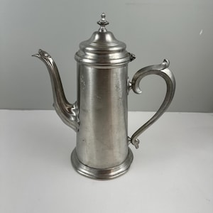 May include: A silver-colored metal coffee pot with a curved handle and a spout. The pot has a lid with a decorative finial. The design is vintage and classic, suitable for serving coffee or tea. The pot is made of a shiny, reflective metal.