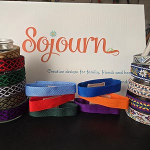 May include: A collection of fabric ribbons and straps featuring various woven patterns and solid colors. The items are arranged around two glass bottles, with a white sign in the background displaying the text Sojourn Creative designs for family, friends and home.