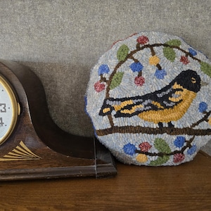 May include: A round, hand-stitched pillow featuring a yellow and black bird perched on a branch with blue and red berries. The bird is set against a light blue background. The pillow is placed next to a wooden clock.