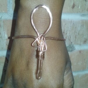 May include: A copper wire hand ornament featuring a central ankh-like symbol with a looped top, a triangular center, and a wrapped base that extends down the back of the hand, accompanied by a matching copper wire ring with a double spiral design.