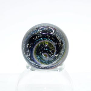 May include: A glass paperweight with a swirling galaxy design in shades of blue, green, and black. The paperweight is on a clear glass stand.