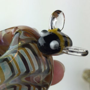 May include: A glass figurine of a bee with a striped yellow and black body, clear wings, and black eyes. The bee is sitting on a hand and is made of clear glass with a swirling pattern of brown, white, and blue.