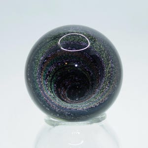 May include: A black glass marble with a swirling, galaxy-like pattern in shades of purple, green, and gold.