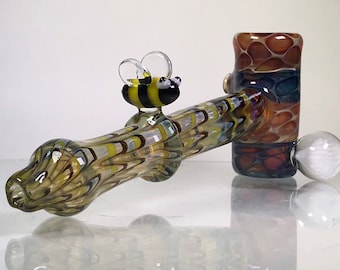 May include: A glass pipe with a bee design on the mouthpiece and a clear glass bowl. The pipe is decorated with a swirl pattern of brown, yellow, and blue glass.