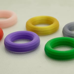 May include: A collection of six colorful, ring-shaped objects. The rings are in shades of red, pink, yellow, purple, green, and gray. Each ring has a textured, ridged surface, and they are arranged on a white surface.