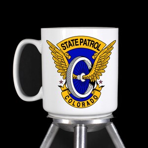 May include: White ceramic coffee mug with a blue and gold design. The design features a winged wheel with the text "STATE PATROL COLORADO".