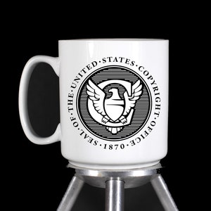 May include: White ceramic mug featuring the Seal of the United States Copyright Office. The black and white seal includes an eagle, wings, and the text "THE SEAL OF THE UNITED STATES COPYRIGHT OFFICE 1870". The mug has a curved handle.