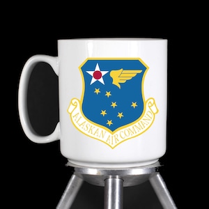 May include: White ceramic mug with a blue and yellow Alaskan Air Command emblem. The emblem features a stylized wing and stars.