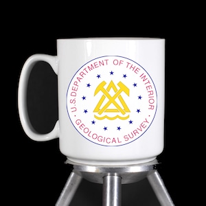 May include: White ceramic mug with a blue and red circle design featuring the text "U.S. DEPARTMENT OF THE INTERIOR. GEOLOGICAL SURVEY." and a gold emblem of crossed pickaxes and a triangle.