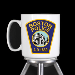May include: White ceramic mug featuring the Boston Police Department emblem. The emblem is a blue shield with gold trim, the words "BOSTON POLICE" in gold, and a building illustration. The text "A.D. 1630" is below the emblem.