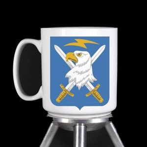 May include: White ceramic mug with a blue and gold design featuring a bald eagle with crossed swords and a lightning bolt. The mug is on a silver stand.