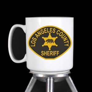 May include: White ceramic mug with a black and gold Los Angeles County Sheriff badge design. The badge features a five-pointed star with a bear inside and the words "Los Angeles County Sheriff".