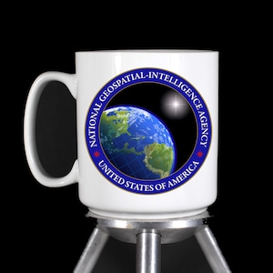 May include: White ceramic mug with a blue and gold logo of the National Geospatial-Intelligence Agency. The logo features a globe with a grid pattern and a star in the background.