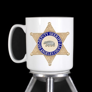 May include: White ceramic mug with a gold and blue Los Angeles County Sheriff's Department star badge. The badge features a bear and the words "Deputy Sheriff Los Angeles County".