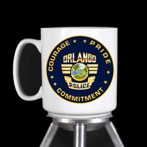 May include: White ceramic mug with a blue and gold Orlando Police Department logo. The logo features a winged shield with the words "Orlando Police" and a depiction of a lake. The words "Courage, Pride, Commitment" are written around the logo.
