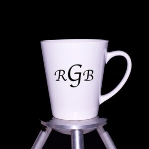 Puede incluir: Taza de cer&aacute;mica blanca con la inscripci&oacute;n "RGB" en negro, sentada sobre un tr&iacute;pode plateado sobre un fondo negro.
