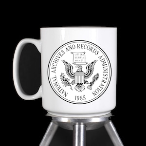 May include: White ceramic mug with a black and white image of the National Archives and Records Administration seal. The seal features an eagle with a scroll that reads "Littera Scripta Manet" and the year 1985.