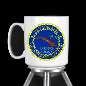 May include: White ceramic mug with a blue and yellow seal depicting the US Naval Base Guantanamo Bay, Cuba. The seal features a chain, a star, and the words "US Naval Base Guantanamo Bay, Cuba".