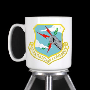 May include: White ceramic mug with a blue and yellow graphic of the Strategic Air Command logo. The logo features a stylized arm holding a lightning bolt and a branch of leaves.