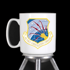 May include: White ceramic mug with a blue and gold Air Force Communications Command logo. The logo features a globe with lightning bolts and stars.