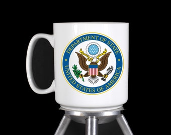 May include: White ceramic mug with a blue and gold seal of the United States Department of State. The seal features an eagle with a shield and olive branches.
