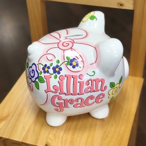 May include: White ceramic piggy bank with a pink bow and floral design. The name "Lillian Grace" is written in pink letters.