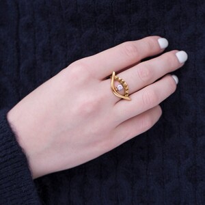 May include: A gold-colored ring with an eye-shaped design. The ring features a central, light-colored gemstone and smaller, amber-colored stones along the top curve. The ring is worn on a hand with white nail polish, set against a dark blue sweater.