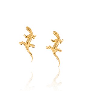 Puede incluir: Un par de pendientes dorados con forma de lagarto. Cada pendiente presenta un diseño texturizado detallado, con el cuerpo, las patas y la cola del lagarto claramente definidos. Los pendientes se muestran sobre un fondo blanco, con un sutil reflejo.
