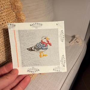 May include: A square needlepoint canvas featuring a seagull wearing a red scarf. The design is printed on white mesh with a vertical orange line on the left. The text Saltbox Needlepoint Cohasset, MA is printed along the edges of the canvas.