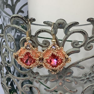 May include: A pair of handcrafted earrings with a square, faceted, pink crystal center. The earrings are surrounded by peach-colored beads and smaller gold-colored beads. Gold-colored ear wires complete the design. The earrings are displayed on a decorative metal stand.