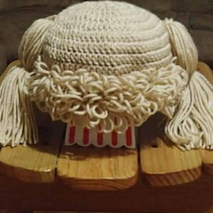May include: A handmade, off-white crochet hat with long yarn strands resembling hair. The hat features a textured crown and a fringe of looped yarn around the brim. The hat is displayed on a wooden stand with a red and white striped design.