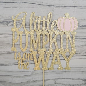 Puede incluir: Adorno dorado con purpurina para pastel con la frase "A little PUMPKIN is on the WAY" y una ilustraci&oacute;n de calabaza rosa. El adorno est&aacute; sujeto a un palito de madera.