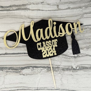 Graduation cap shape glitter cake topper personalized with name across the top and Class of 2024 along the bottom. Shown here with the name Madison in gold glitter on a black graduation cap.