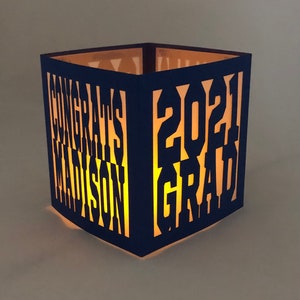 May include: A navy blue, square lantern with cut-out text that reads "CONGRATS MADISON 2021 GRAD." The lantern is illuminated from within, creating a warm glow. A unique graduation gift, ideal for a celebration.