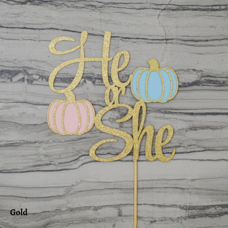 May include: A gold glitter cake topper with the words "He or She" and two pumpkin illustrations, one pink and one blue. The topper is attached to a wooden stick for easy insertion into a cake. The text is in a cursive font.