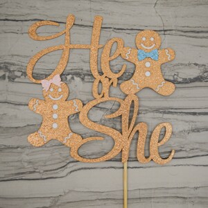 May include: A glittery gold cake topper with the words "He or She" in cursive script. Two gingerbread people, one with a pink bow and the other with a blue bow tie, flank the text. The topper is attached to a wooden stick.