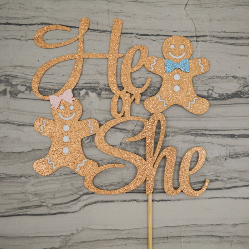 May include: A glittery gold cake topper with the words "He or She" in cursive script. Two gingerbread people, one with a pink bow and the other with a blue bow tie, flank the text. The topper is attached to a wooden stick.