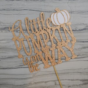 Puede incluir: Un adorno para pastel dorado con purpurina con la frase "A little PUMPKIN is on the WAY" y una calabaza blanca. El adorno est&aacute; sujeto a un palito de madera para insertarlo en el pastel.
