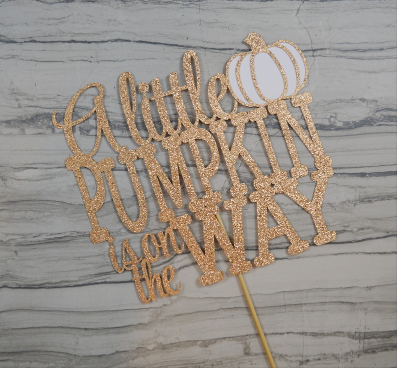 Puede incluir: Un adorno para pastel dorado con purpurina con la frase "A little PUMPKIN is on the WAY" y una calabaza blanca. El adorno est&aacute; sujeto a un palito de madera para insertarlo en el pastel.